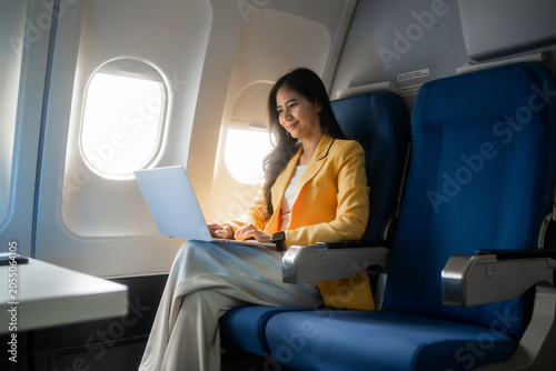 Young woman using a laptop while sitting by the window on an airplane, enjoying a calm flight. Ideal for travel, business, remote work, tourism, and modern lifestyle themes.