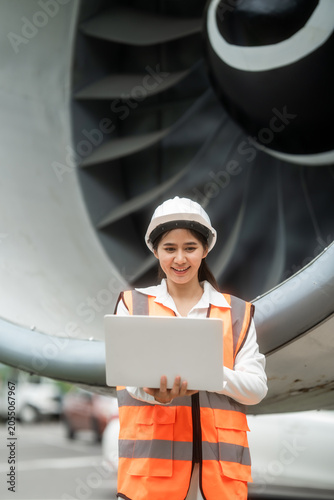 Female airport ground engineer in hard hat and safety vest using a laptop beside a large airplane engine, showing aircraft inspection, maintenance, and aviation operations.