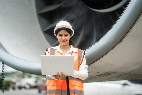 Female airport ground engineer in hard hat and safety vest using a laptop beside a large airplane engine, showing aircraft inspection, maintenance, and aviation operations.