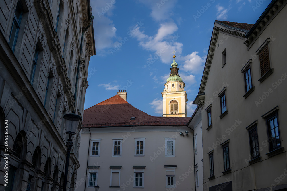 Obraz premium Narrow Old Town passage framing church tower and traditional buildings in Ljubljana showcasing European heritage and street photography charm