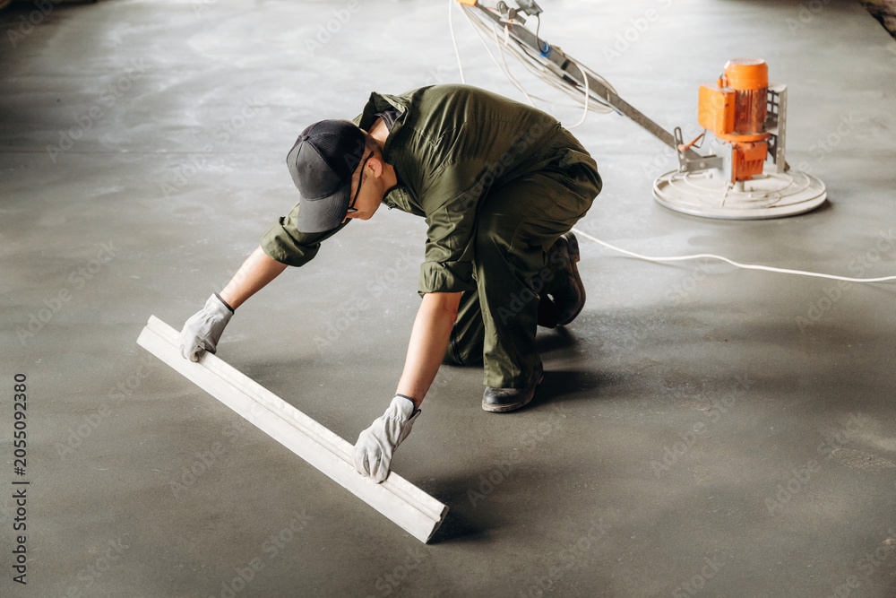 Naklejka premium Male construction worker in green coveralls uses a leveling tool on freshly poured concrete floor with a power trowel visible in the background