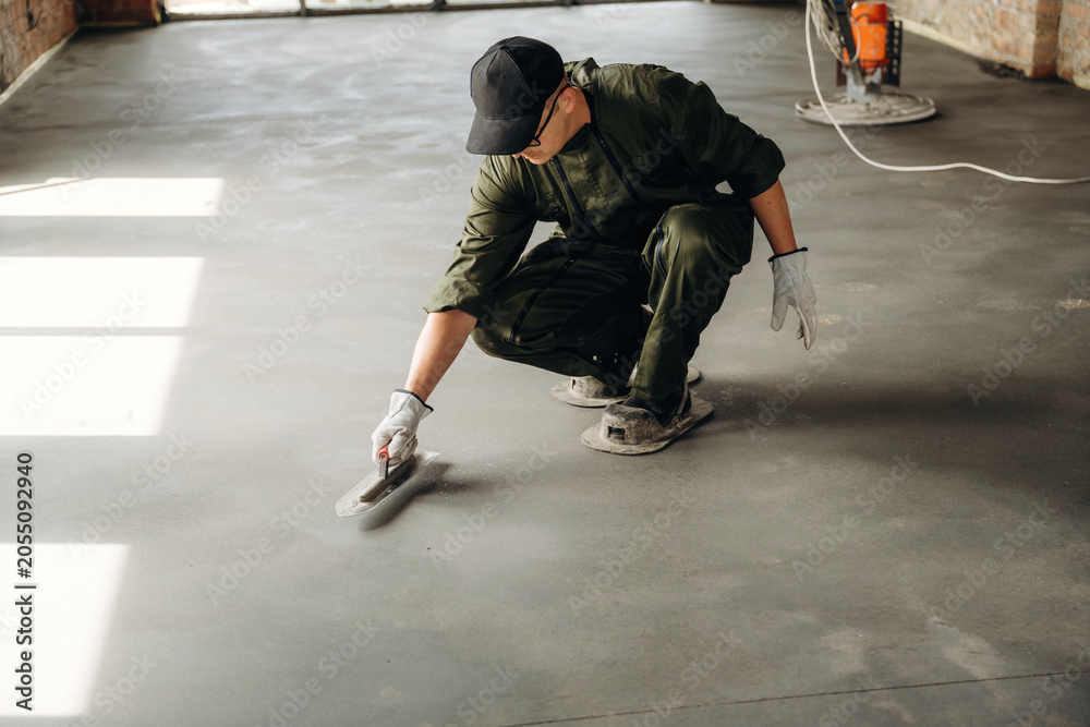 Naklejka premium Male construction worker in green coveralls kneeling on a concrete floor applying finishing touches with a trowel in an industrial setting with natural light