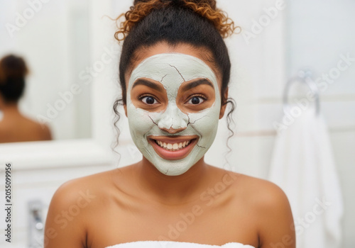 Young woman applying face mask and smiling in bathroom mirror  