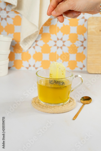 Hand dropping tea bag into a glass cup of hot tea on a white table