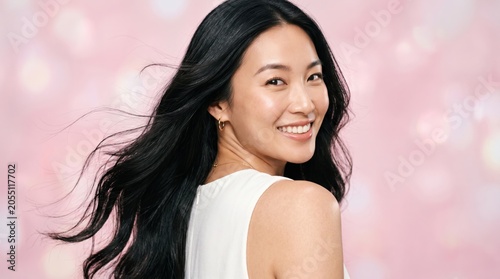 Beautiful Asian woman smiling with flowing long black hair looking over her shoulder. Glow skin, clean beauty makeup, white dress, soft pink bokeh background for skincare, hair care, cosmetics promo