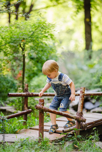 Blond toddler boy in denim overalls walking on wooden bridge among green plants in summer garden.