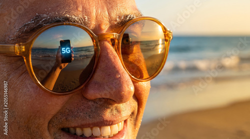 Close-up of man with 5G smartphone screen reflected in his round sunglasses.