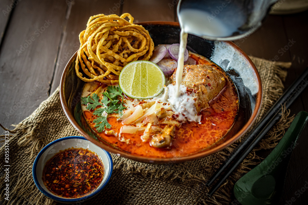 Poster Action shot of pouring soup into a traditional bowl of Northern Thai Khao Soi chicken curry noodle with crispy egg noodles and lime, served on a dark rustic wooden background table