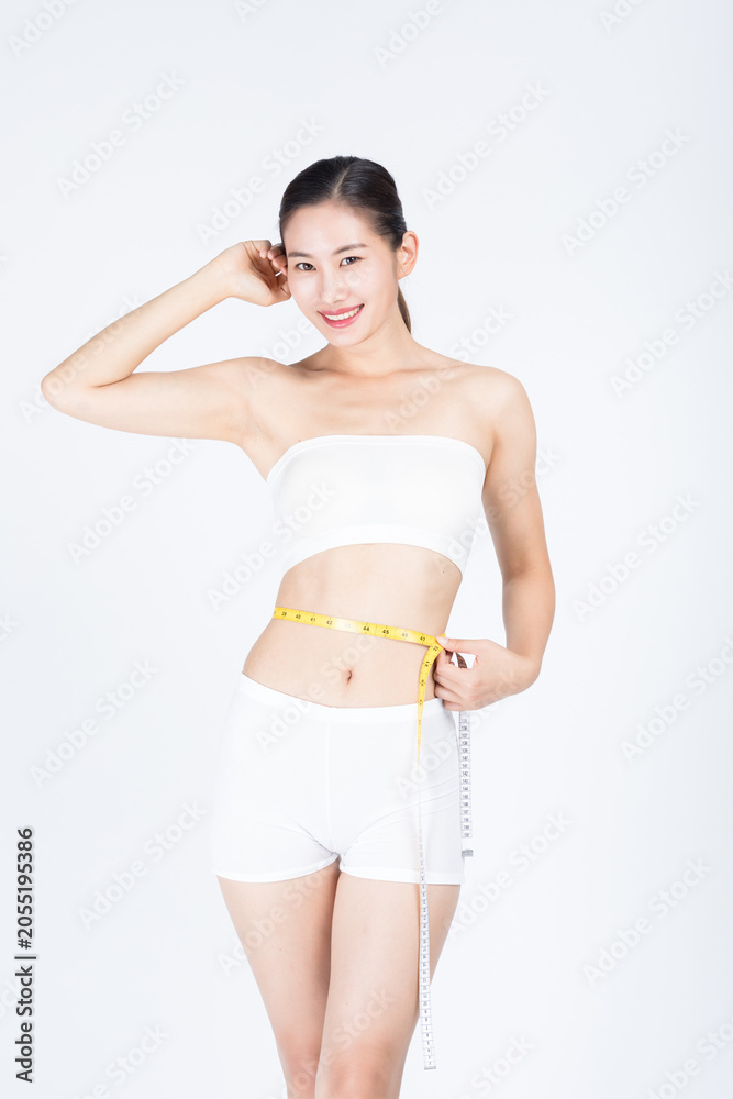 Fototapeta premium Smiling young woman measuring waist with tape measure in white activewear
