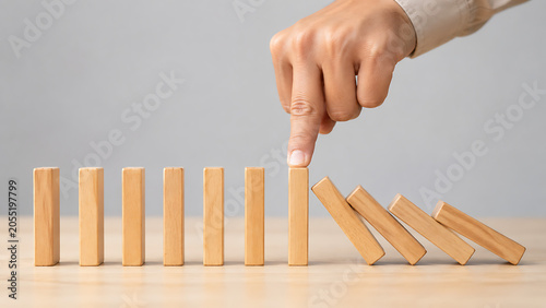 Hand stops the domino effect by pressing down on a light wooden block preventing the chain reaction of falling pieces in crisis management