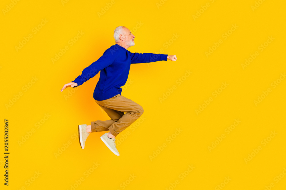 Naklejka premium Active senior man jumping joyfully against a bright yellow background wearing a blue sweater and casual sneakers for fitness, energy and positive lifestyle imagery