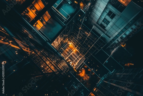 Tilted view of tall city buildings with glowing lights and reflections at night, creating a moody urban scene with strong lines and modern architecture.