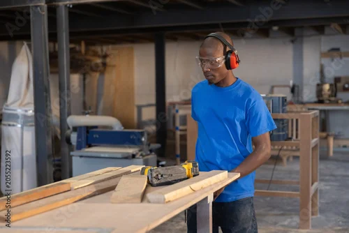 Fototapeta Carpenter is measuring wood planks on workbench in spacious workshop with machines around