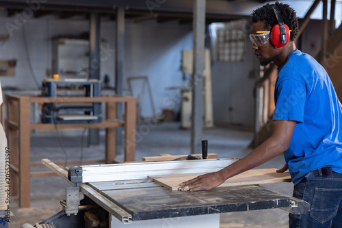 Table saw blade is slicing wooden plank on workbench in industrial shop with dust-extraction hose