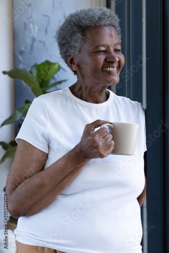 Senior African woman standing at home by dark doorframe wearing white ribbed tee holding beige mug