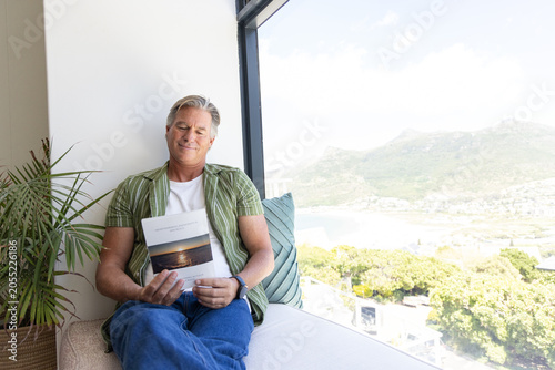Fotografie Senior man sitting on cushioned window bench holding sunset booklet, leaning aga