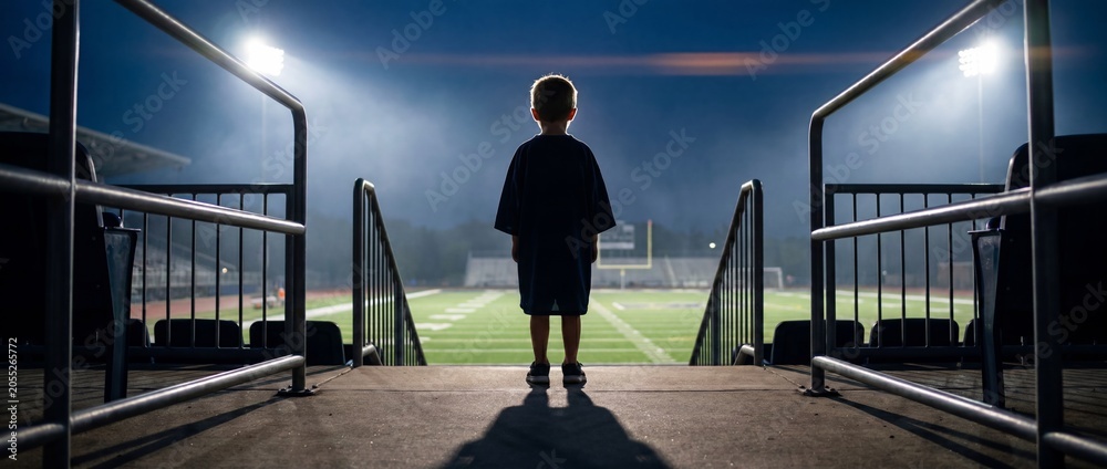 Naklejka premium Young boy looking at empty american football field under stadium lights. Concept of childhood dream, future athlete, sports ambition, inspiration and family fan culture. Banner