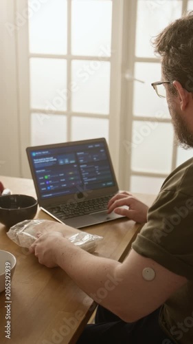 Vertical shot of Caucasian young adult man with glucose sensor checking food package nutrition and typing on laptop using health dashboard. For diabetes care and medical tech marketing