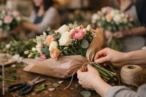 Close up of hands wrapping fresh flower bouquet with twine and scissors on table, handmade floral arrangement preparation