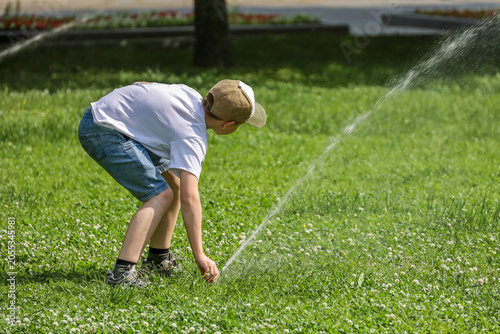 Kid boy plays with water jets on a lawn. Child enjoying the summer, hot weather in city park