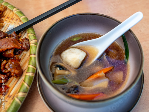 Asian soup with mushroom and vegetable in bowl on plate on table. Part of plate with chicken piece in soy sauce.