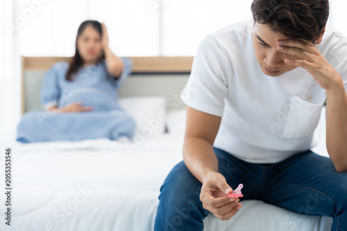 Man holding condom package while pregnant woman sits in background, representing family planning, responsibility, and lifestyle concept in cozy indoor bedroom environment with natural daylight