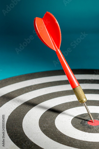 Canvas Print A vertical close-up shot of a single red dart firmly embedded in the red center bullseye of a dartboard against a blue background, representing ultimate success, perfect focus, and definitive victory