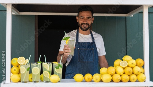 Man in a denim apron smiles while holding a glass of refreshing lemonade at a stand
