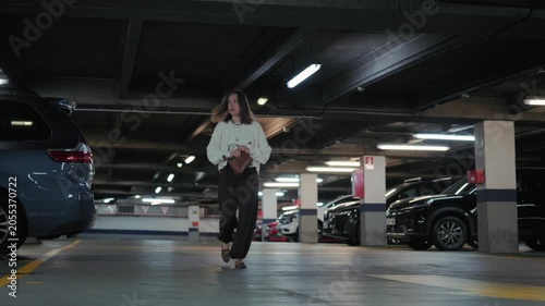 A young woman walks away from her car in an underground parking lot, putting her keys into her handbag as she heads towards the exit.