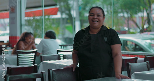 Latina waitress smiling warmly while standing in dining area, hand resting on chair in casual restaurant setting with guests in soft background