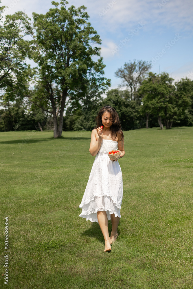 Obraz premium Girl in white dress holds strawberry and wants to eat it. Fragile Asian woman on picnic in park at dawn, summer vacation and outdoor recreation