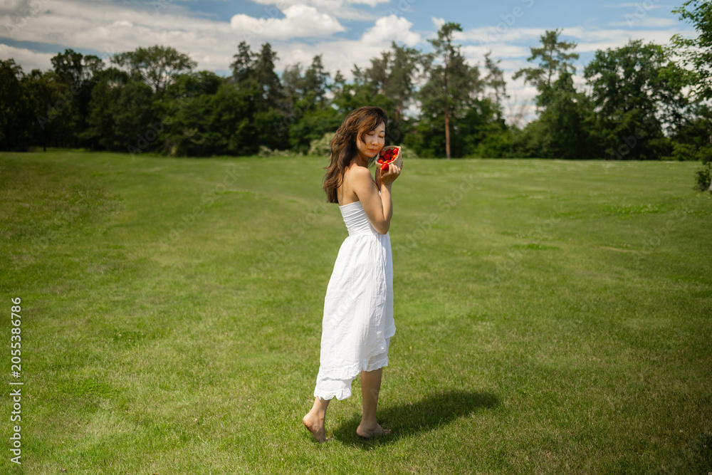 Obraz premium Girl in white dress holds strawberry and wants to eat it. Fragile Asian woman on picnic in park at dawn, summer vacation and outdoor recreation