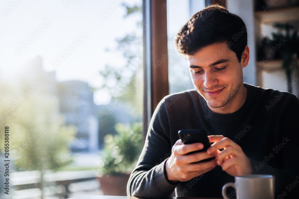 Fototapeta premium Man using smartphone in a coffee shop, smiling and relaxed in natural light