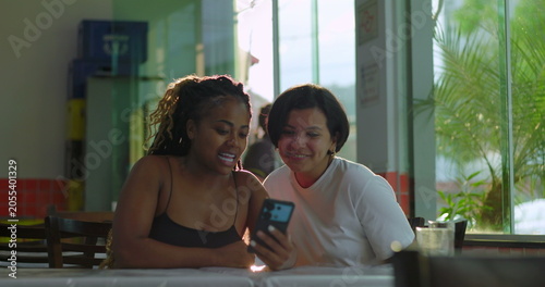 Foto Two women seated at restaurant table react to something on smartphone, smiling a