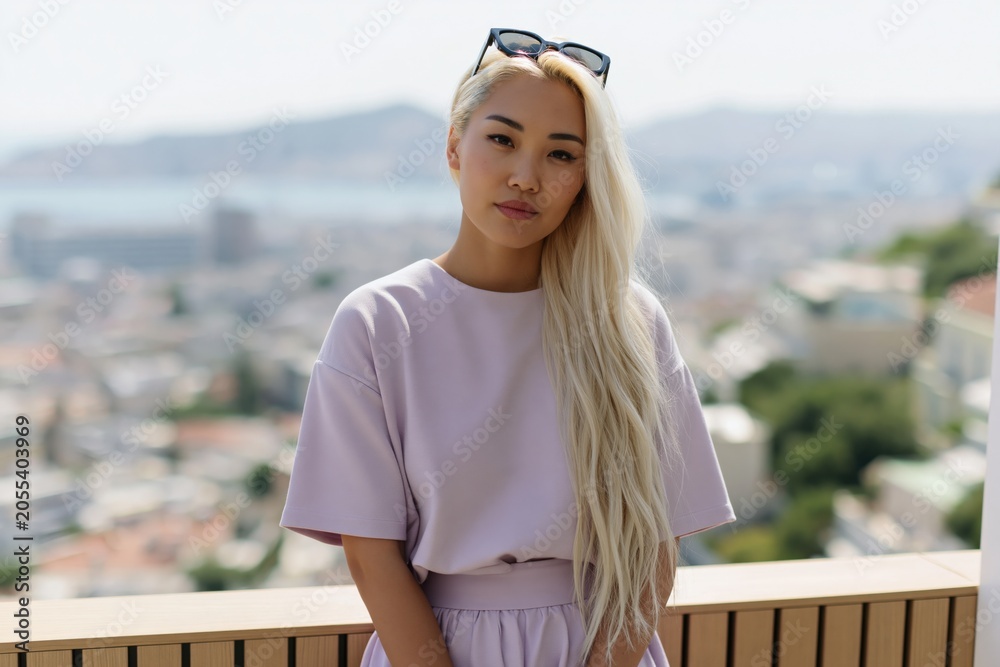 Fototapeta premium Young woman with long platinum hair in pastel top on balcony with blurred cityscape for lifestyle portrait and travel background use