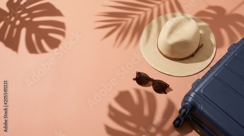 Blue suitcase, straw hat, and sunglasses on pastel peach background with palm leaves shadow. Flat lay summer travel concept with copy space.