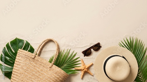 Summer beach flat lay with straw bag, hat, sunglasses, starfish, and palm leaves on white sand background. Tropical vacation concept with copy space.