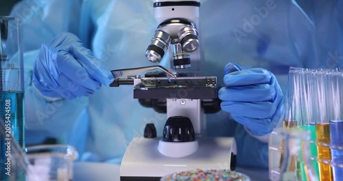 Technician places microplastic particles onto microscope slide in lab setting. Assistant in protective suit analyzes polymer materials in laboratory