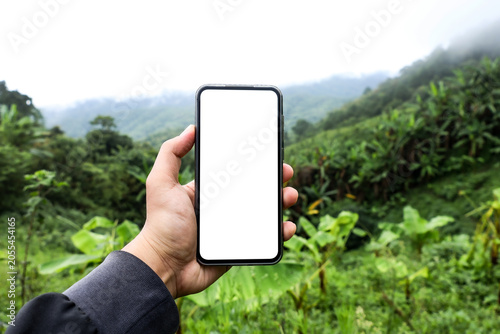 Smartphone mockup with blank white screen held by hand in green nature mountain background.