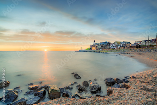The iconic lighthouse in Urk stands sentinel over a tranquil waterfront as the sun dips below the horizon, casting a warm, golden glow across the calm sea and sky.