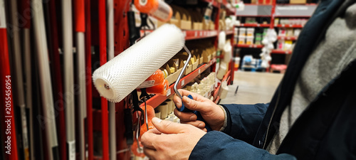 A customer holds a paint tray and brushes while shopping in a hardware store aisle. Concepts of home improvement, DIY renovation, consumer choice, and retail shopping behavior.