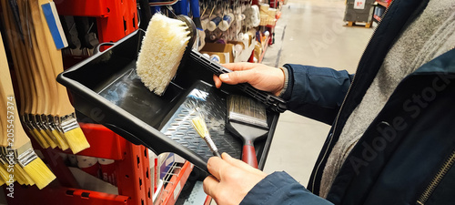 A customer holds a paint tray and brushes while shopping in a hardware store aisle. Concepts of home improvement, DIY renovation, consumer choice, and retail shopping behavior.
