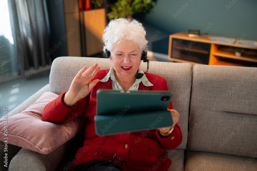 Obraz premium A senior woman sits on a couch, wearing a headset and smiling while using a tablet for a video call. She waves her hand, engaged in conversation, in a cozy living room.