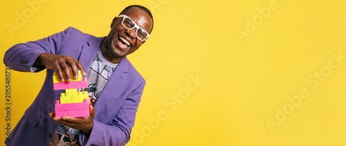Happy black man playing blocks in purple suit with colorful toy building bricks laughing joyfully against bright yellow background banner copy space for business creativity success strategies concept.