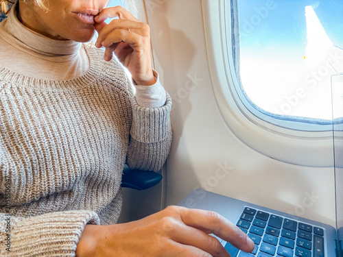 Woman working on a laptop in an airplane window seat, thoughtful and focused during a business flight; typing, remote work and travel lifestyle captured in a cozy sweater and natural daylight.