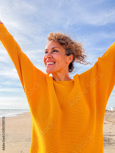 Woman in yellow sweater raising her arms on a sunny beach, smiling with joy and freedom, enjoying outdoors and a carefree moment