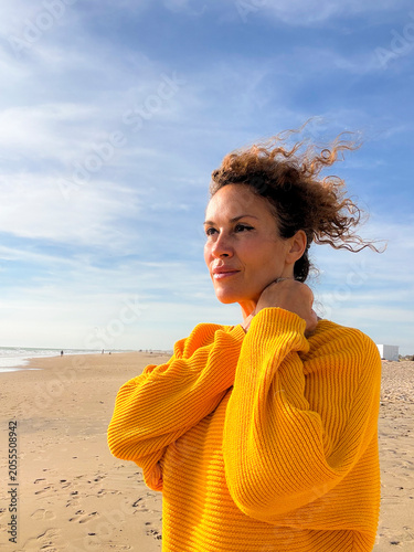 Woman in a yellow sweater standing on a windy beach, portrait of a contemplative middle-aged woman looking toward the horizon in warm sunlight, casual fashion and peaceful coastal scene.