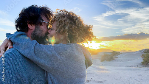 Couple embracing and kissing at sunset on sandy dunes, woman with curly hair and man with beard share intimate moment against colorful sky and warm light