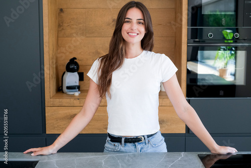 Smiling young woman in white t-shirt leaning on a kitchen counter in a modern home kitchen, casual portrait conveying confidence, relaxation and domestic lifestyle