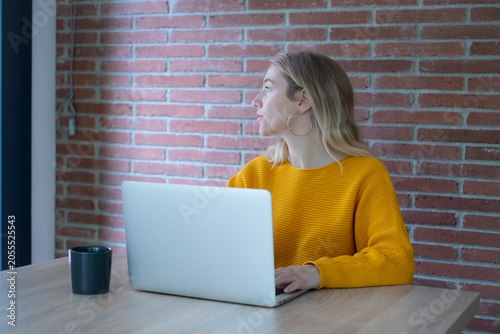 Billede på lærred Young woman in a yellow sweater works on a laptop at a wooden table by a brick wall, looking thoughtfully out the window with a coffee cup nearby in a modern casual workspace
